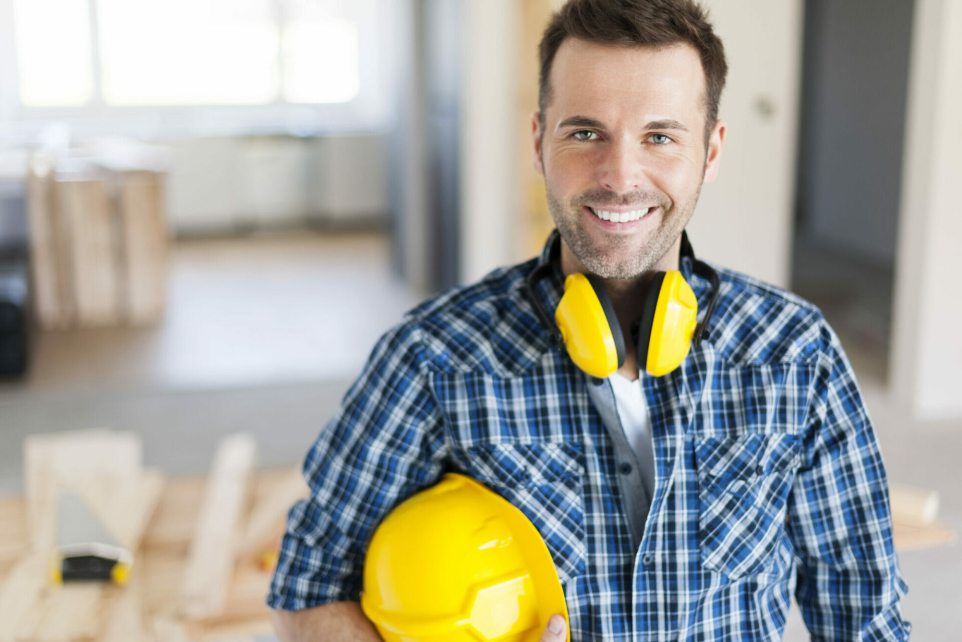 un homme en tenue de chantier souriant tient un casque de chantier et un pair de casque sur ses épaules